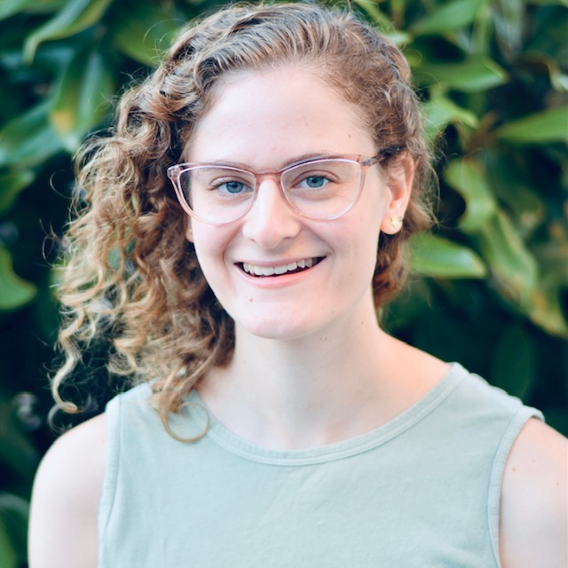 A person with curly, shoulder-length hair, glasses, and a sleeveless shirt smiles in front of a backdrop of green leaves, radiating the same enthusiasm they bring to their lacrosse matches.