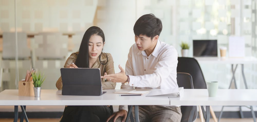 Two people sitting at a desk in an office setting, engaged in a discussion about paid medical leave while looking at a tablet. The person on the left is a woman with long dark hair, and the person on the right is a man with short dark hair. There are plants and office supplies on the desk.